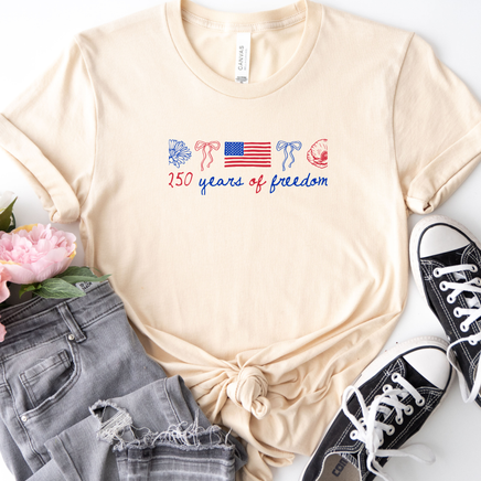 Beige t-shirt with '150 years of freedom' design, paired with black sneakers and gray jeans on a white background.