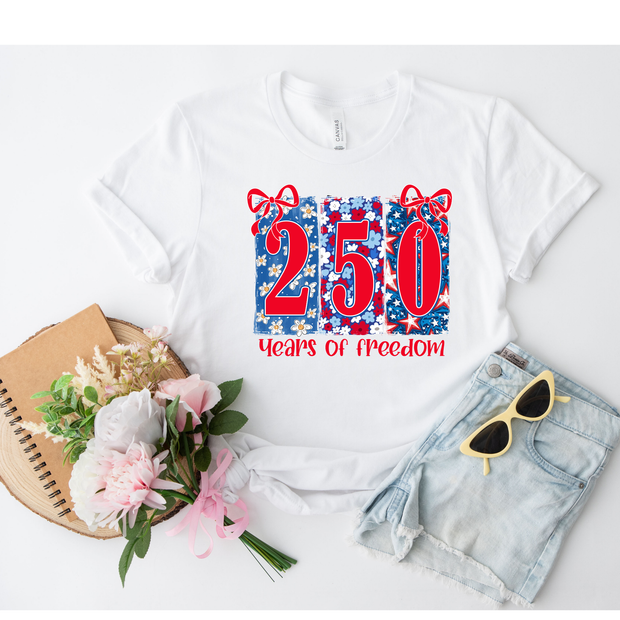 White t-shirt with '250 Years of Freedom' design, sunglasses, denim shorts, flowers, and notebook on a white background